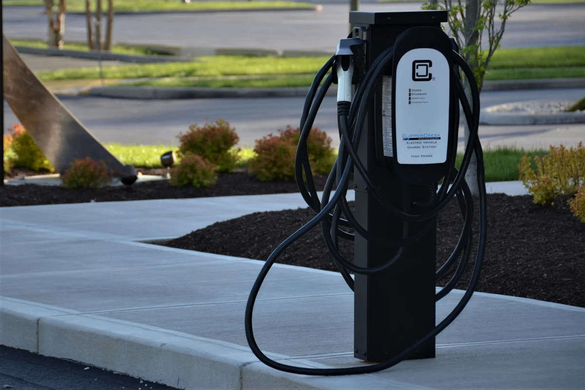 A black electric vehicle charging station with a white face and coiled black cables stands on a concrete sidewalk.