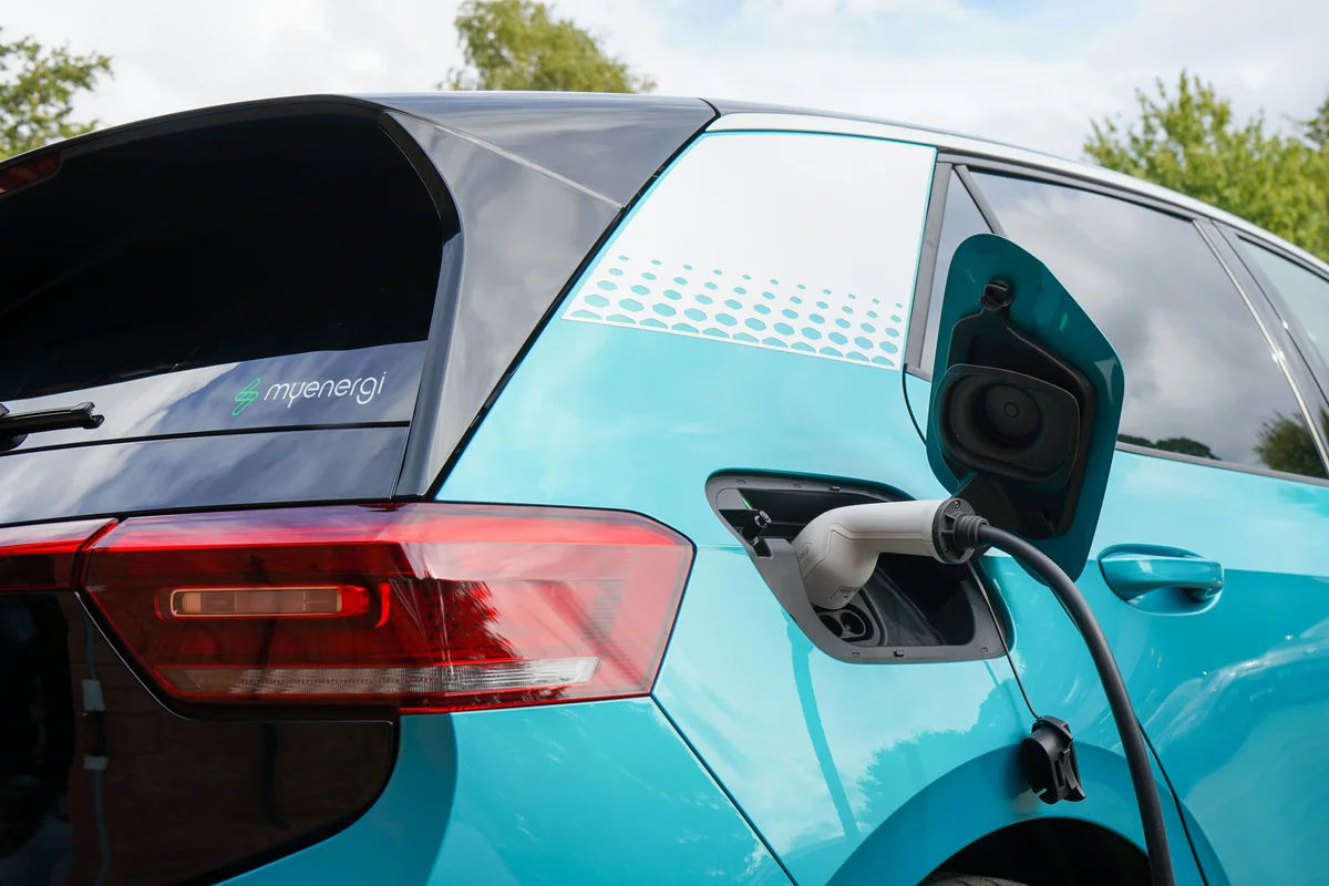A teal electric car is charging, with a white charging cable plugged into its right rear side.