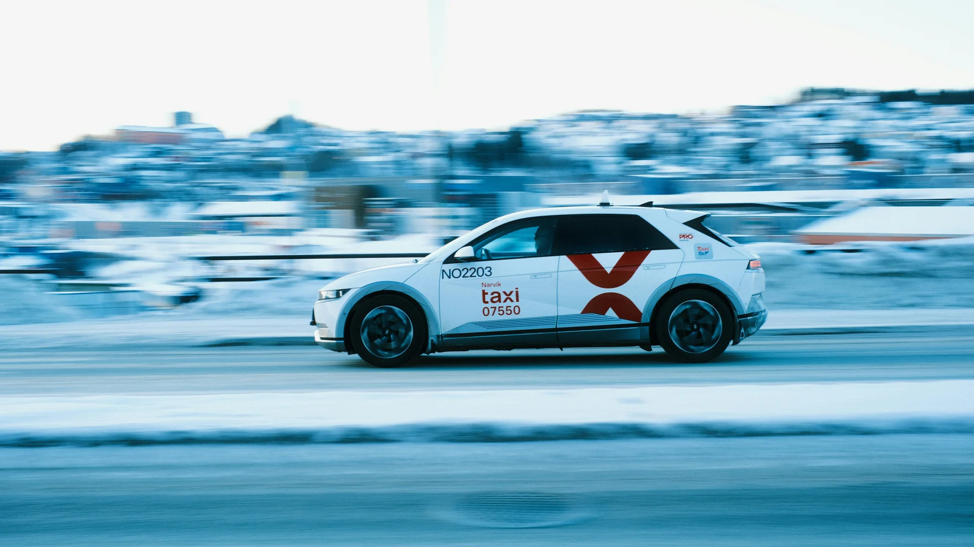 A white taxi with red "taxi" lettering and a large red X on its side drives on a snowy road.