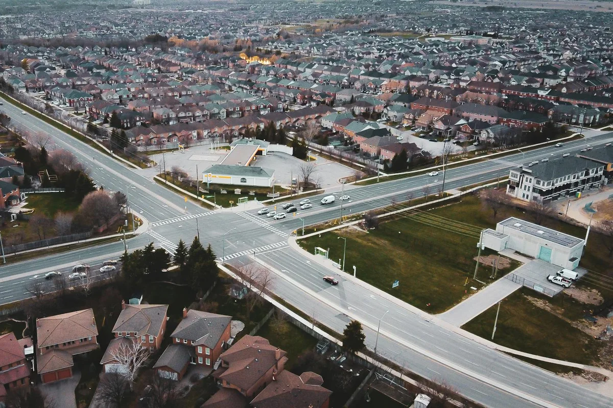 An aerial view of a suburban neighborhood with a large intersection and many houses with dark roofs.