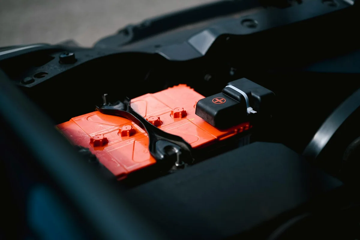 An orange car battery is visible in a dark engine compartment, with a black clamp holding it down.